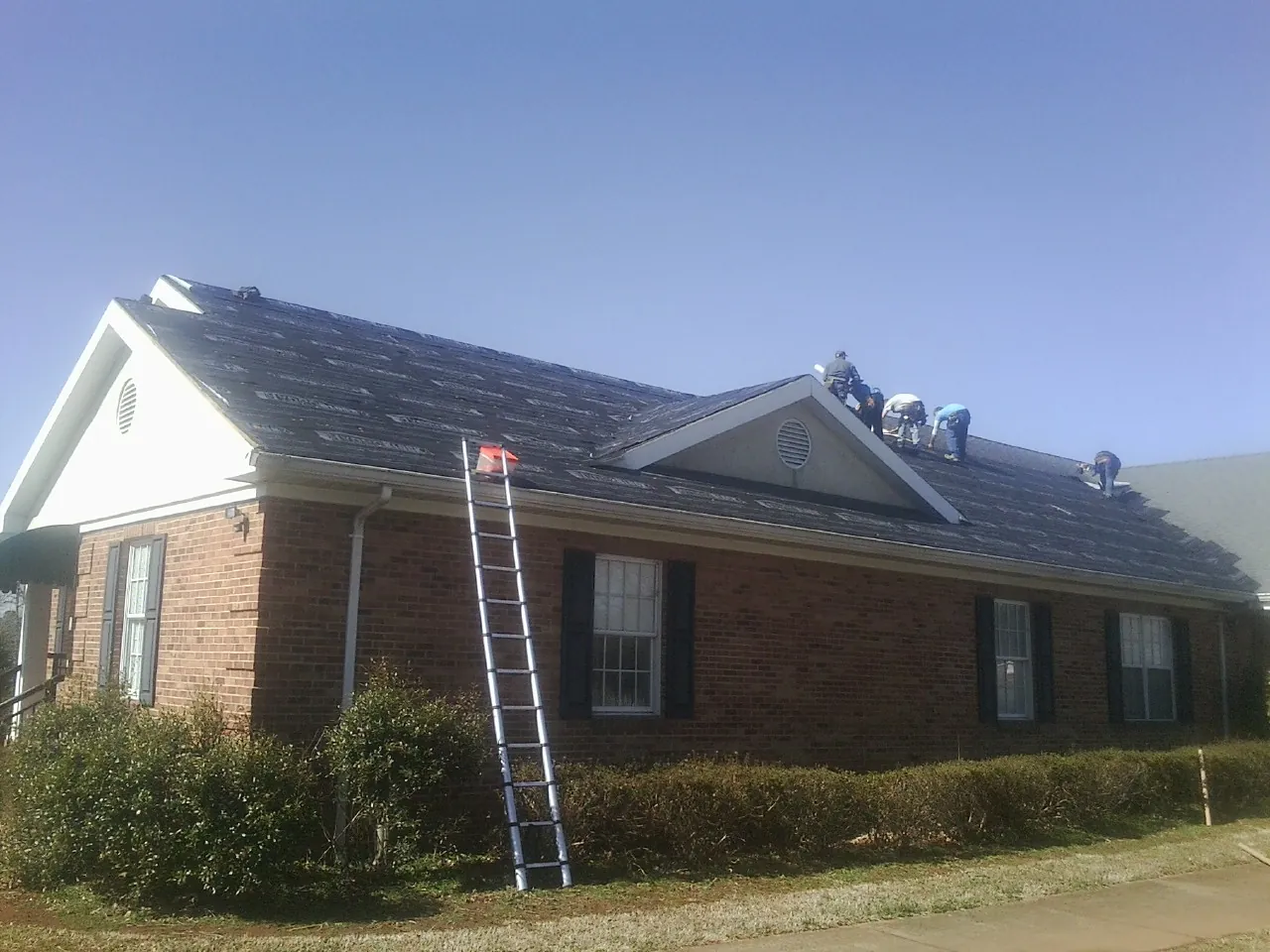 Crew tearing shingles off a brick ranch roof with a ladder set — Emergency Roof Repair work by our crew in Devers